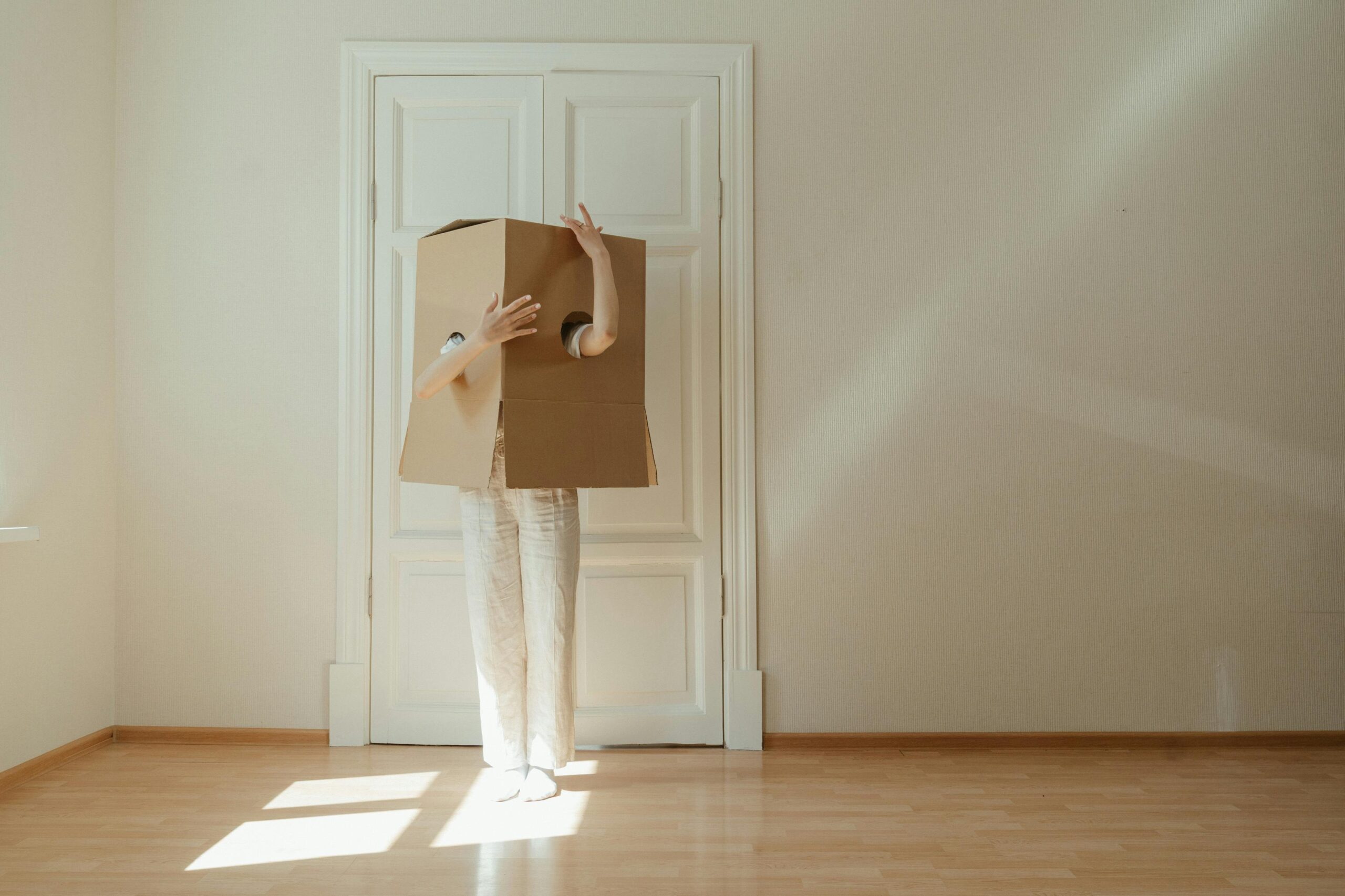 A person stands in a room wearing a cardboard box in a minimalist setting, symbolizing change.